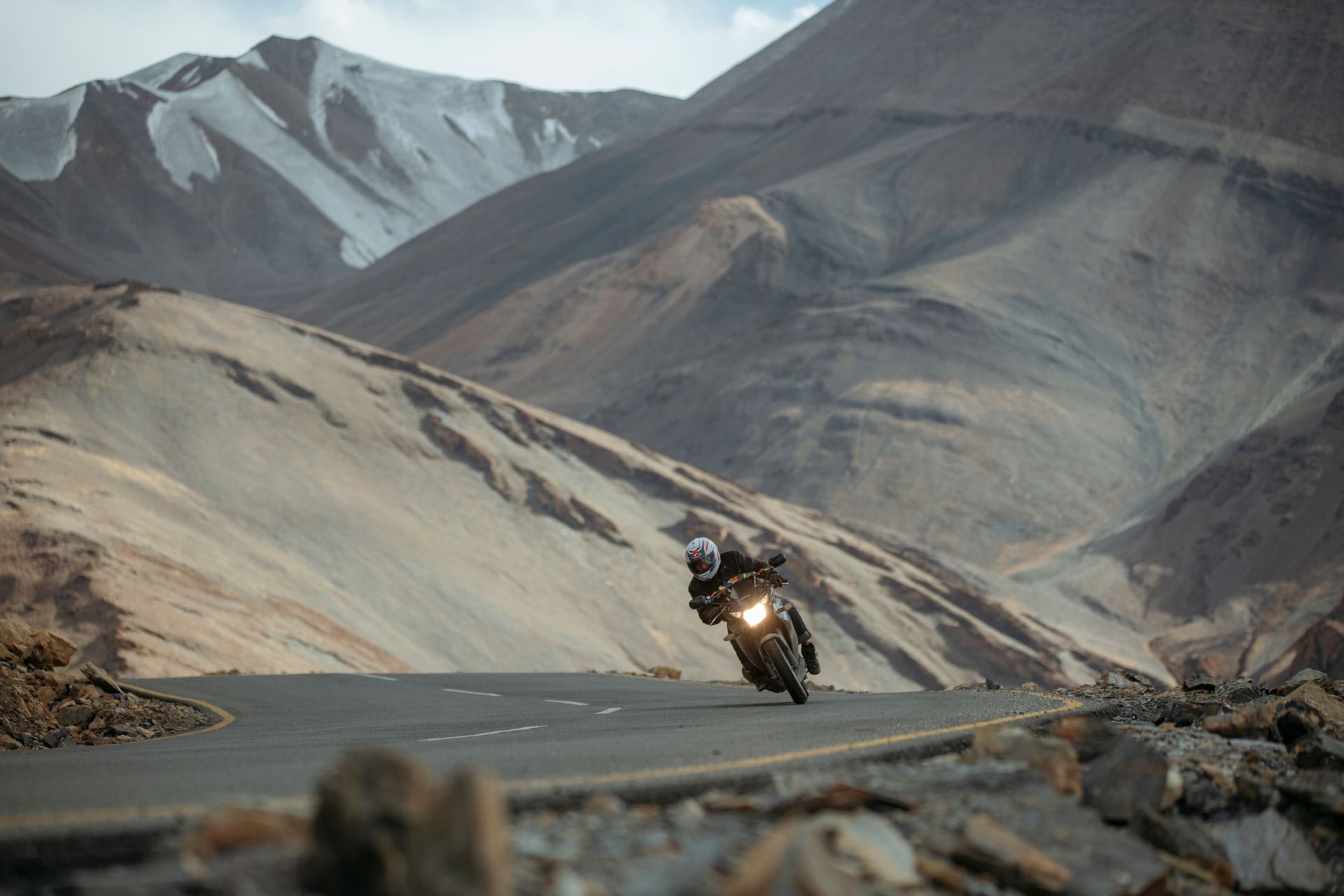 A motorcycle rider leaning into a bend on a high, empty mountain road below snowy peaks